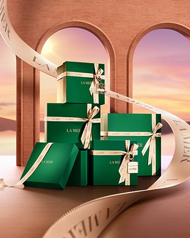 A stack of la mer gift boxes with a larger ribbon running up behind them against a background of arch windows and golden sunset.